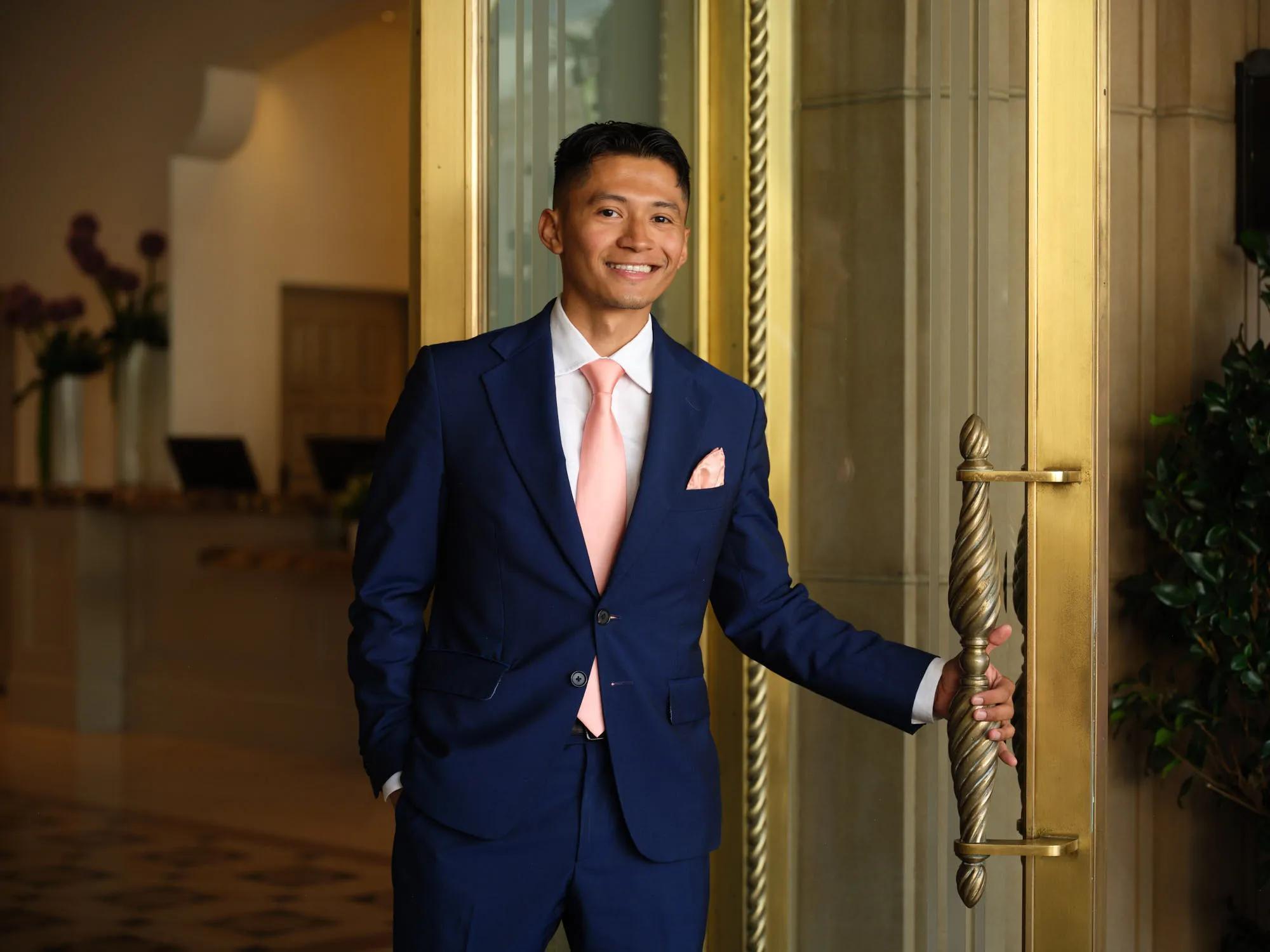 Person in a navy suit with a pink tie opens an ornate gold hotel door, standing in an elegant Beverly Hills lobby.