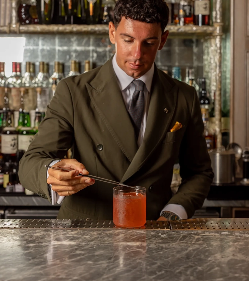 Bartender in a tailored suit stirs an orange cocktail at a polished bar counter.