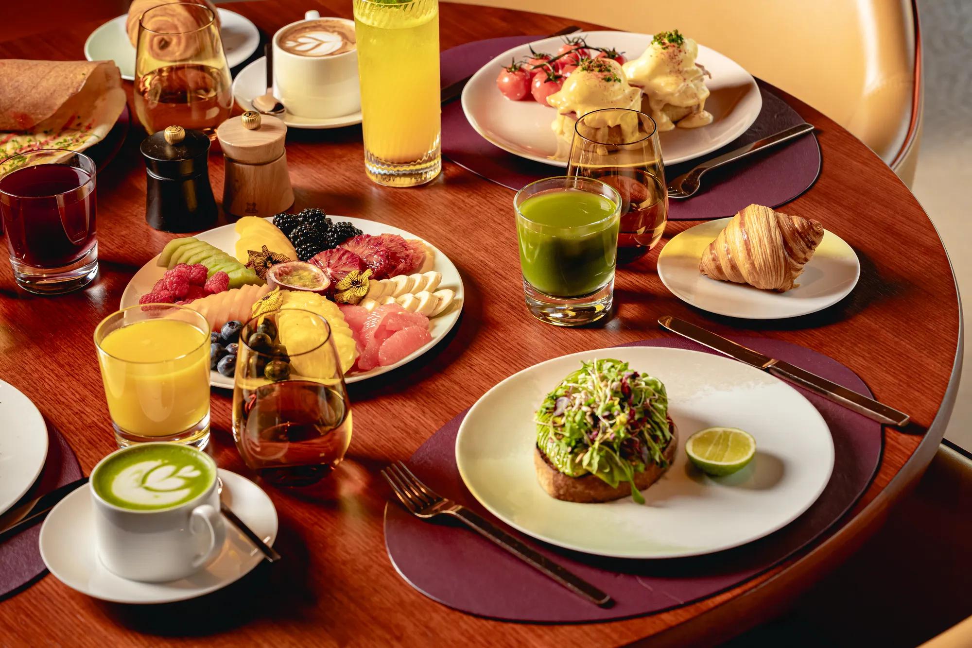 Breakfast table with fruit platter, pastries, avocado toast, eggs Benedict, juices, coffee, and tea on a wooden table.