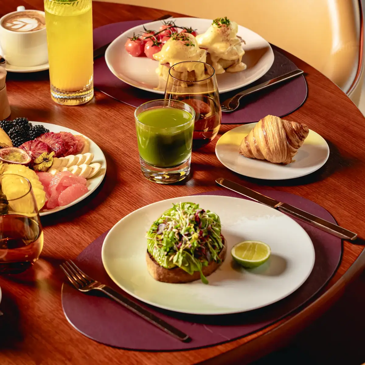Breakfast table with fruit platter, pastries, avocado toast, eggs Benedict, juices, coffee, and tea on a wooden table.