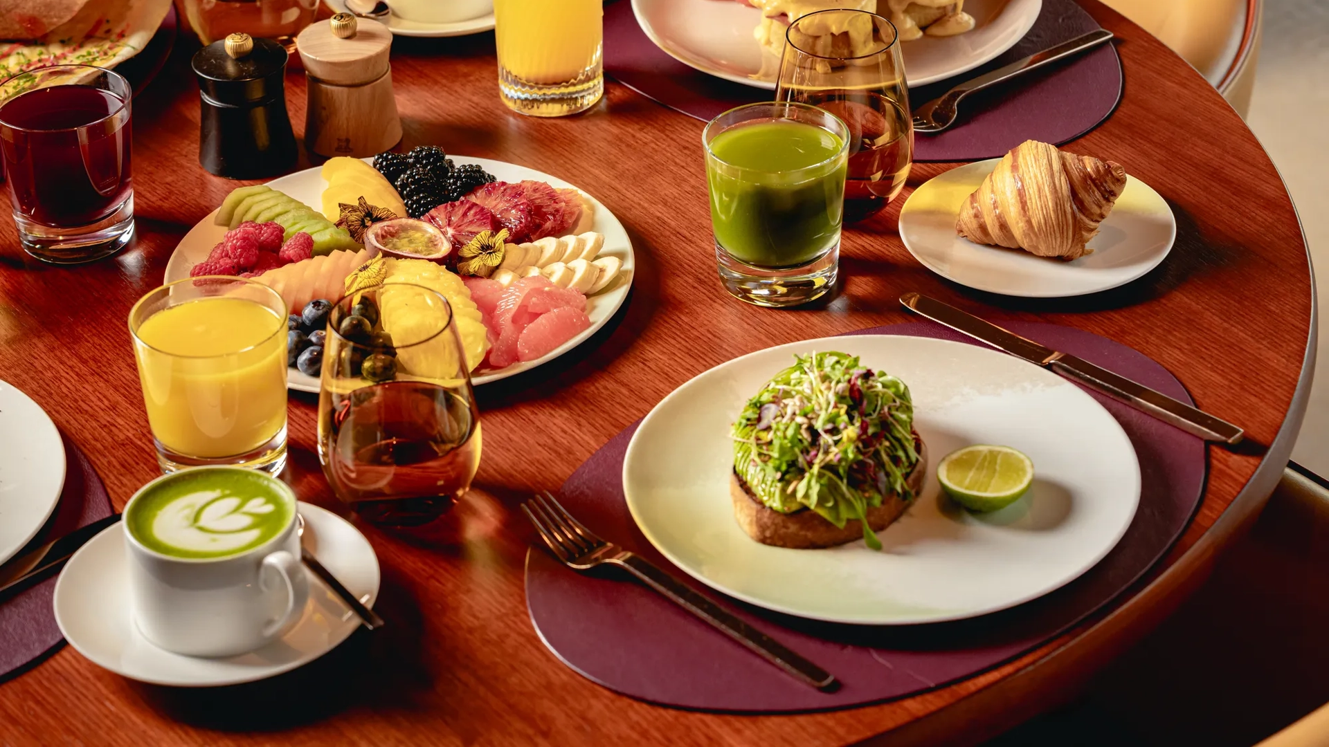 Breakfast table with fruit platter, pastries, avocado toast, eggs Benedict, juices, coffee, and tea on a wooden table.