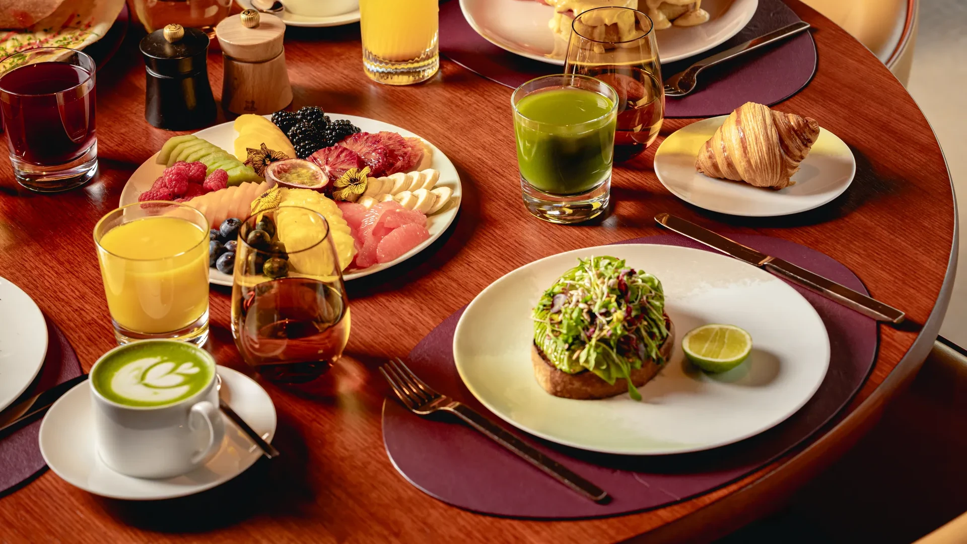 Breakfast table with fruit platter, pastries, avocado toast, eggs Benedict, juices, coffee, and tea on a wooden table.