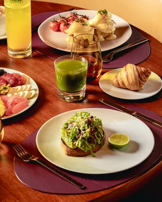 Breakfast table with fruit platter, pastries, avocado toast, eggs Benedict, juices, coffee, and tea on a wooden table.