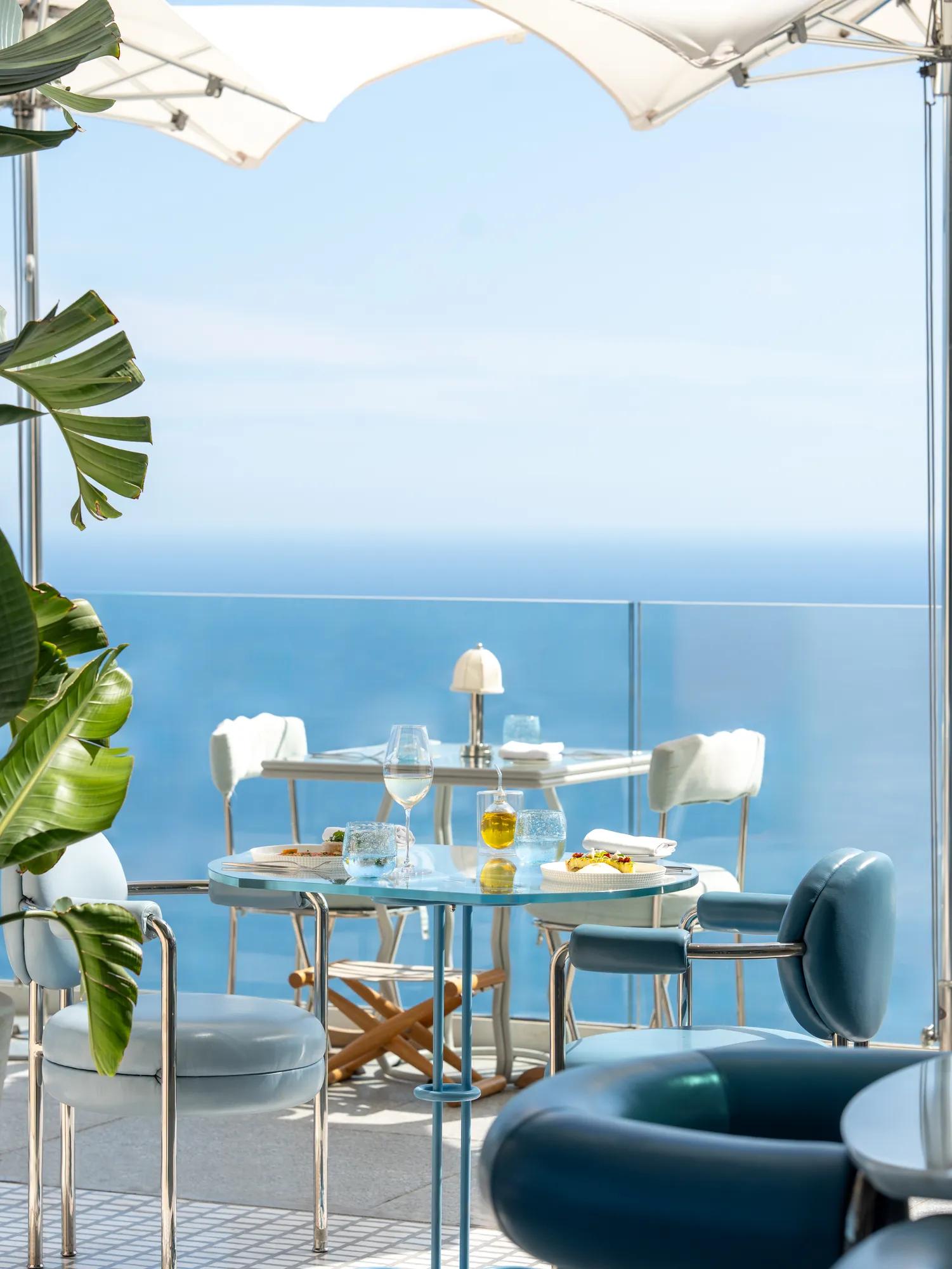 Outdoor dining tables with blue chairs overlooking a clear ocean view under white parasols.