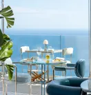 Tables de repas en extérieur avec chaises bleues face à une vue dégagée sur la mer sous des parasols blancs.