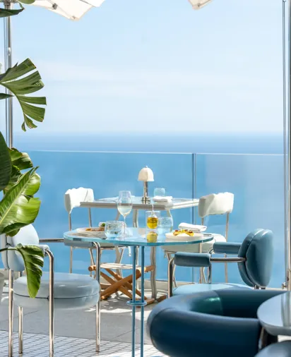 Tables de repas en extérieur avec chaises bleues face à une vue dégagée sur la mer sous des parasols blancs.