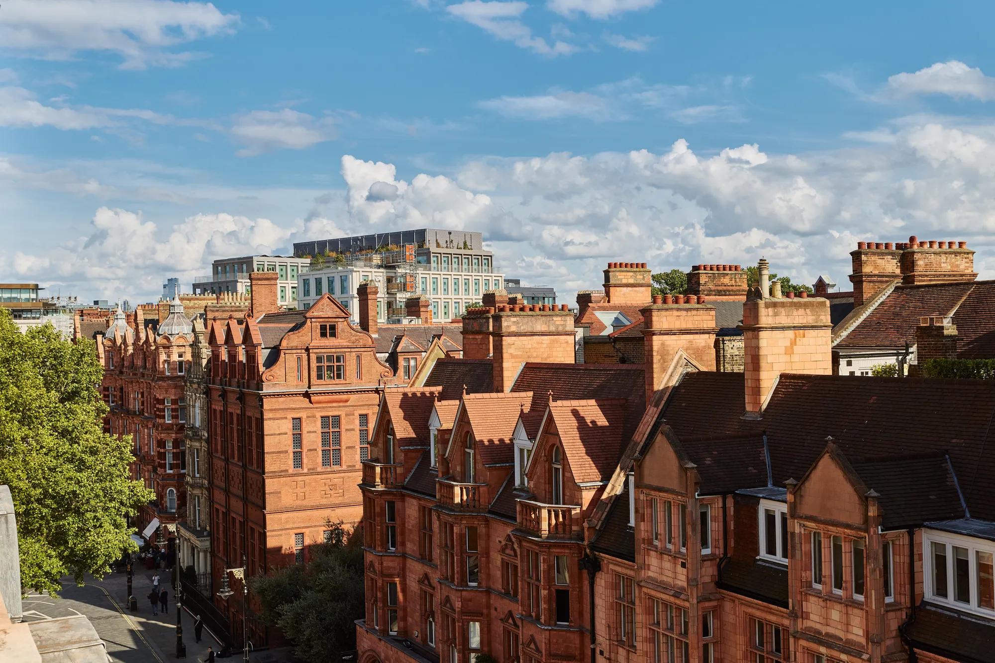 View over red-brick Victorian buildings and chimneys in Mayfair under a bright blue sky with scattered clouds.