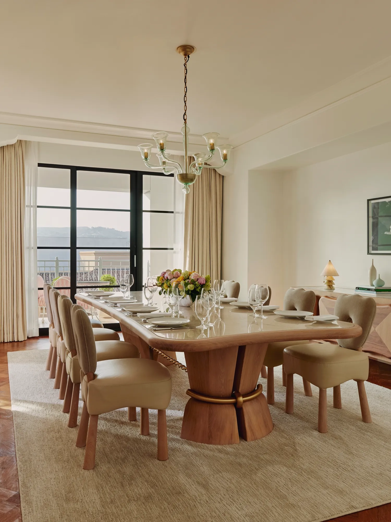 Formal dining room with a long wooden table set for ten, cream upholstered chairs, chandelier, and large windows opening to a balcony with city and hill views.