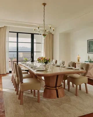 Formal dining room with a long wooden table set for ten, cream upholstered chairs, chandelier, and large windows opening to a balcony with city and hill views.