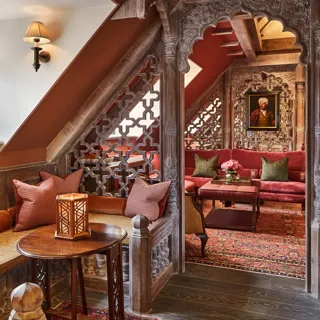 Cosy alcove seating with carved wood screens, red upholstery, patterned rug, and stained glass window in a richly detailed room.