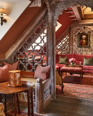 Cosy alcove seating with carved wood screens, red upholstery, patterned rug, and stained glass window in a richly detailed room.