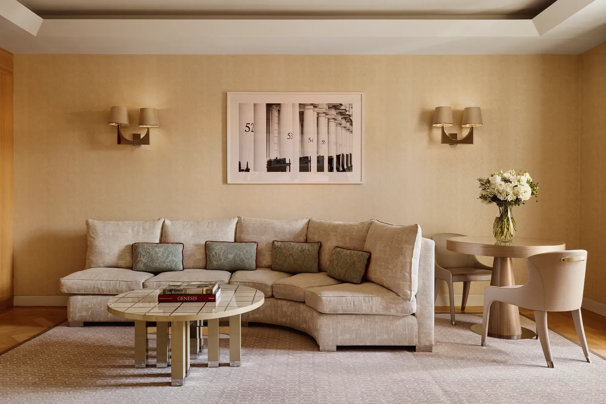 Neutral-toned living room with a curved cream sofa and patterned cushions, round tiled coffee table with books, small dining table with two chairs, and a framed black-and-white artwork on the wall.