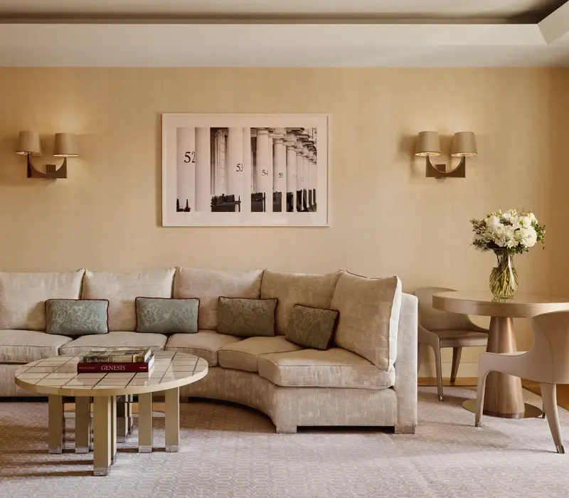 Neutral-toned living room with a curved cream sofa and patterned cushions, round tiled coffee table with books, small dining table with two chairs, and a framed black-and-white artwork on the wall.