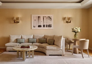 Neutral-toned living room with a curved cream sofa and patterned cushions, round tiled coffee table with books, small dining table with two chairs, and a framed black-and-white artwork on the wall.