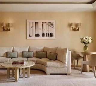 Neutral-toned living room with a curved cream sofa and patterned cushions, round tiled coffee table with books, small dining table with two chairs, and a framed black-and-white artwork on the wall.