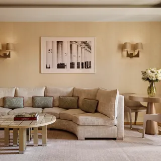 Neutral-toned living room with a curved cream sofa and patterned cushions, round tiled coffee table with books, small dining table with two chairs, and a framed black-and-white artwork on the wall.