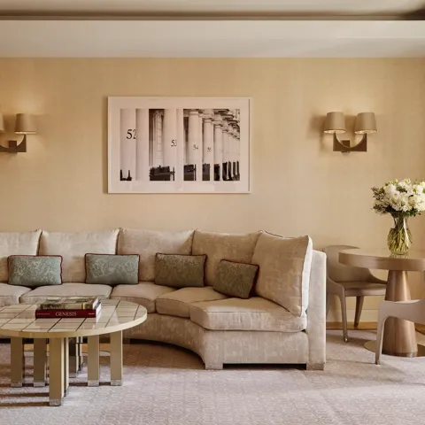 Neutral-toned living room with a curved cream sofa and patterned cushions, round tiled coffee table with books, small dining table with two chairs, and a framed black-and-white artwork on the wall.