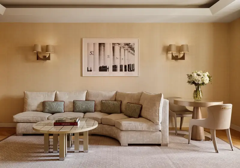 Neutral-toned living room with a curved cream sofa and patterned cushions, round tiled coffee table with books, small dining table with two chairs, and a framed black-and-white artwork on the wall.