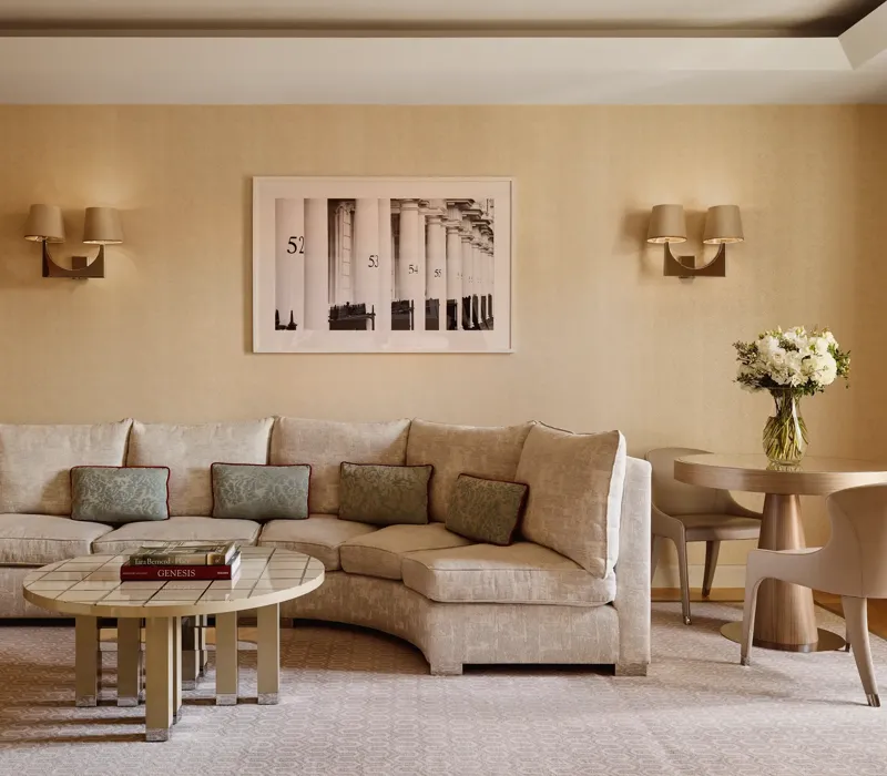 Neutral-toned living room with a curved cream sofa and patterned cushions, round tiled coffee table with books, small dining table with two chairs, and a framed black-and-white artwork on the wall.