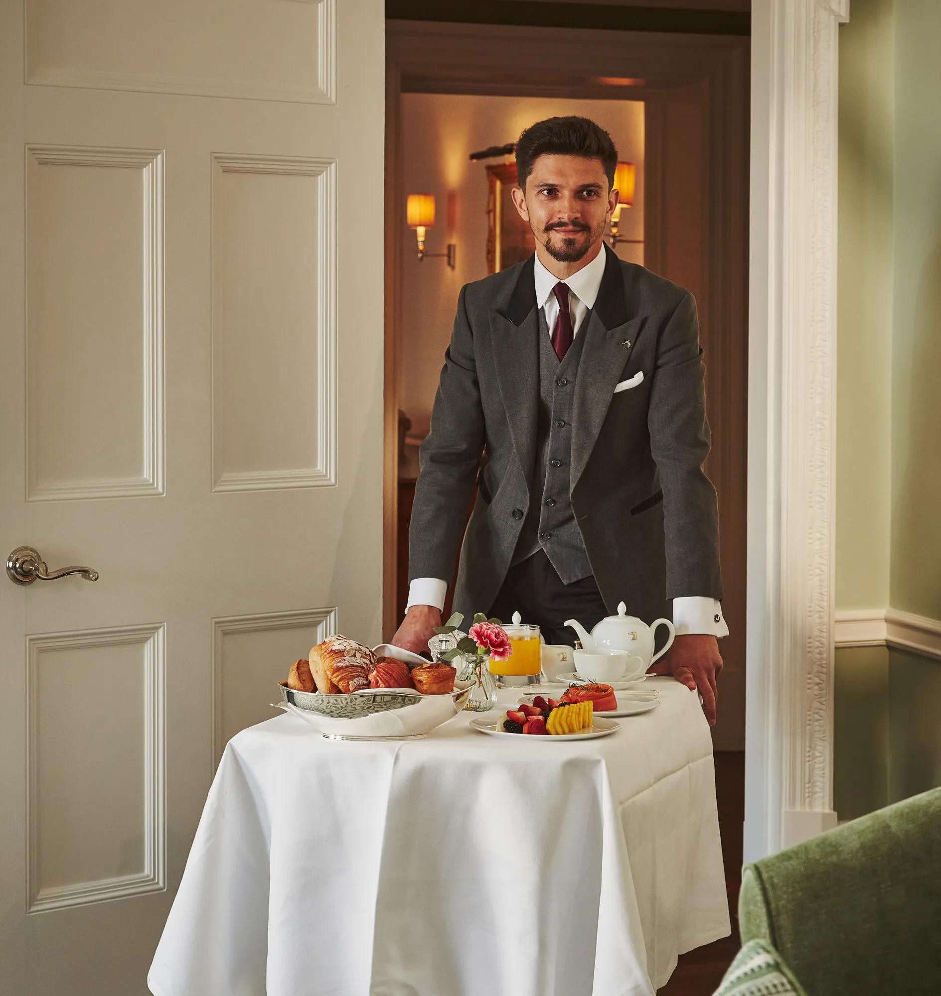 Server in a grey suit presents a breakfast trolley with pastries, fruit, and tea in a softly lit room.