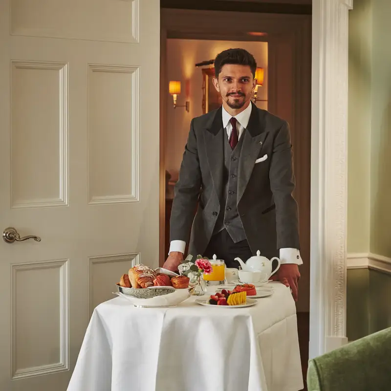 Server in a grey suit presents a breakfast trolley with pastries, fruit, and tea in a softly lit room.