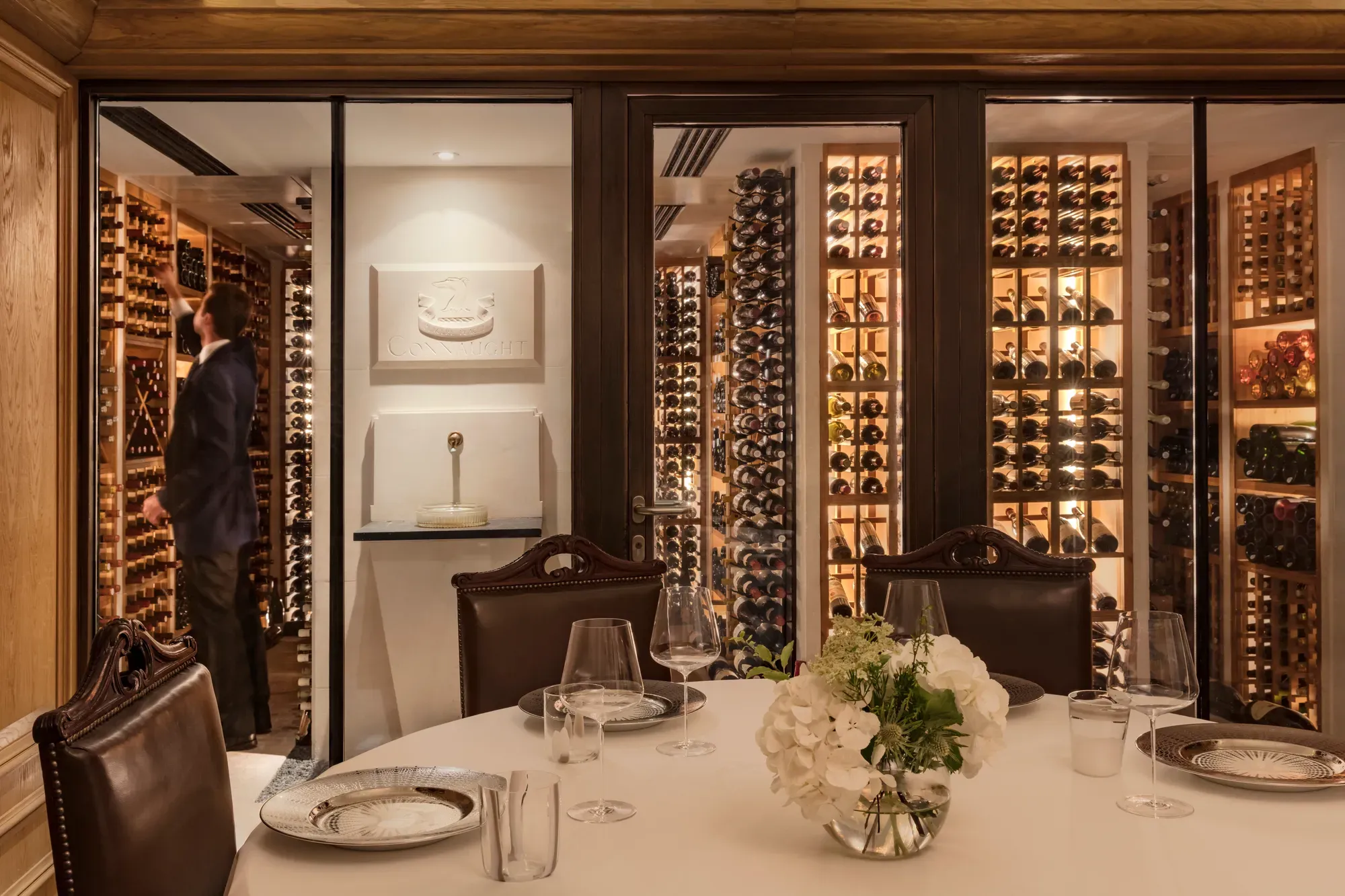 Private dining table with glassware and flowers, set beside a glass-walled wine cellar with shelves of bottles.