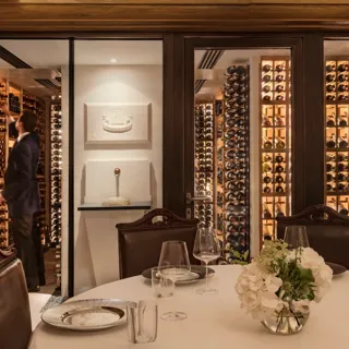 Private dining table with glassware and flowers, set beside a glass-walled wine cellar with shelves of bottles.