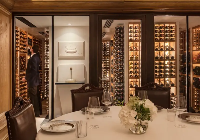 Private dining table with glassware and flowers, set beside a glass-walled wine cellar with shelves of bottles.