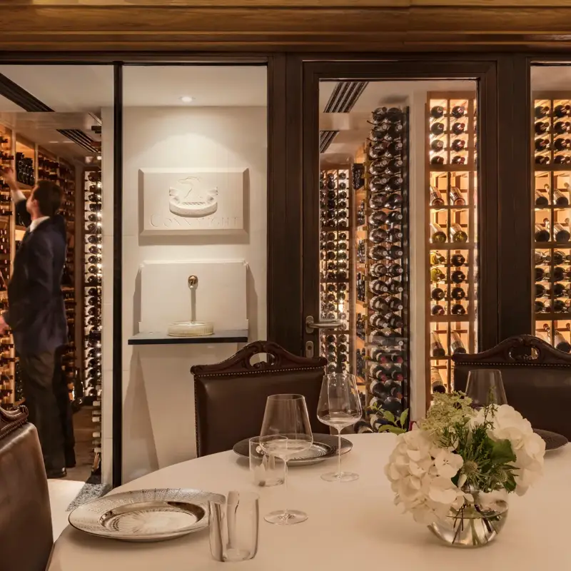 Private dining table with glassware and flowers, set beside a glass-walled wine cellar with shelves of bottles.