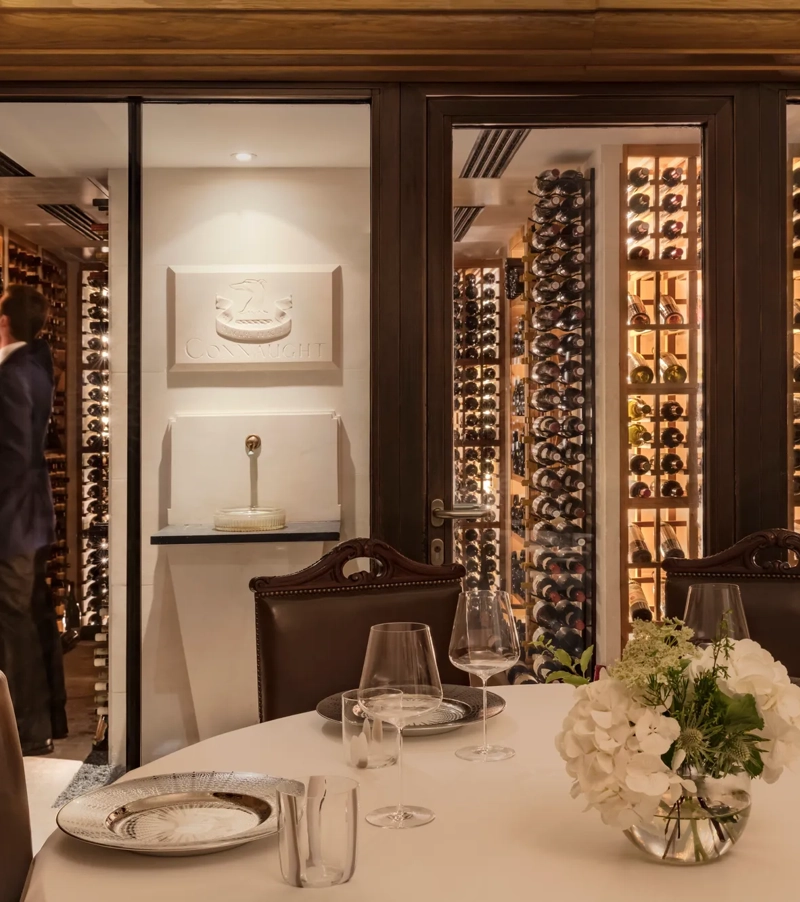Private dining table with glassware and flowers, set beside a glass-walled wine cellar with shelves of bottles.
