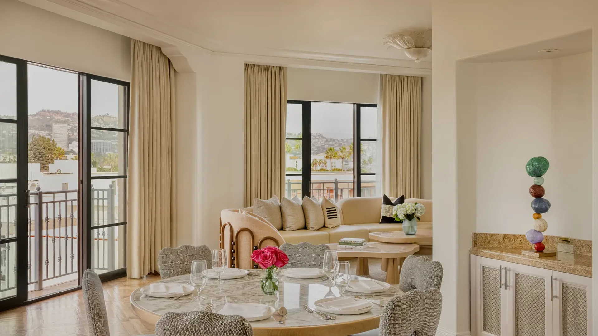 Hollywood Suite dining area with round table, cushioned chairs, with a curved couch in the foreground with Beverly Hills skyline views.