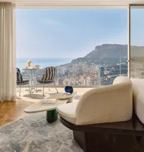 Bright living area opening onto a balcony with bistro seating, overlooking a coastal city and sea beyond.