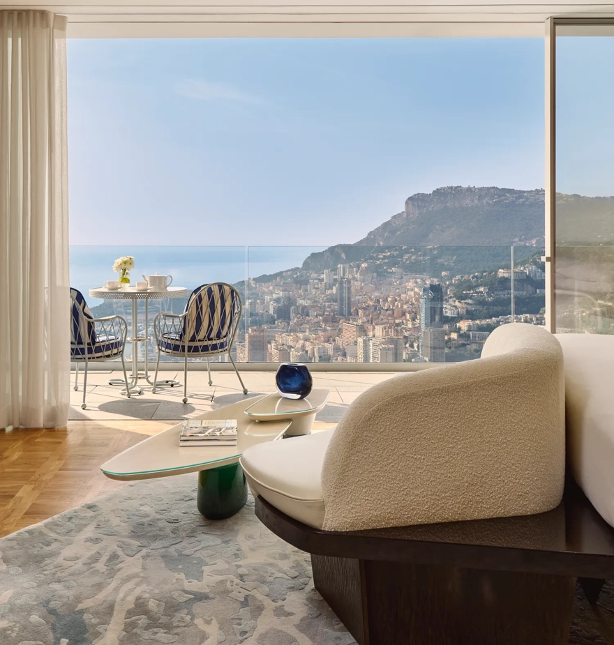 Bright living area opening onto a balcony with bistro seating, overlooking a coastal city and sea beyond.