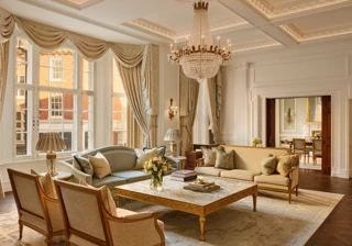 Grand living room with high ceilings, ornate chandelier, and large windows draped in gold curtains, featuring elegant sofas and armchairs around a marble coffee table with carved gold detailing.