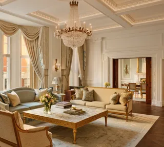 Grand living room with high ceilings, ornate chandelier, and large windows draped in gold curtains, featuring elegant sofas and armchairs around a marble coffee table with carved gold detailing.