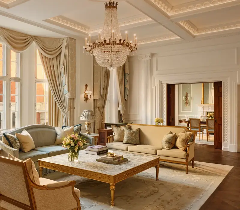Grand living room with high ceilings, ornate chandelier, and large windows draped in gold curtains, featuring elegant sofas and armchairs around a marble coffee table with carved gold detailing.