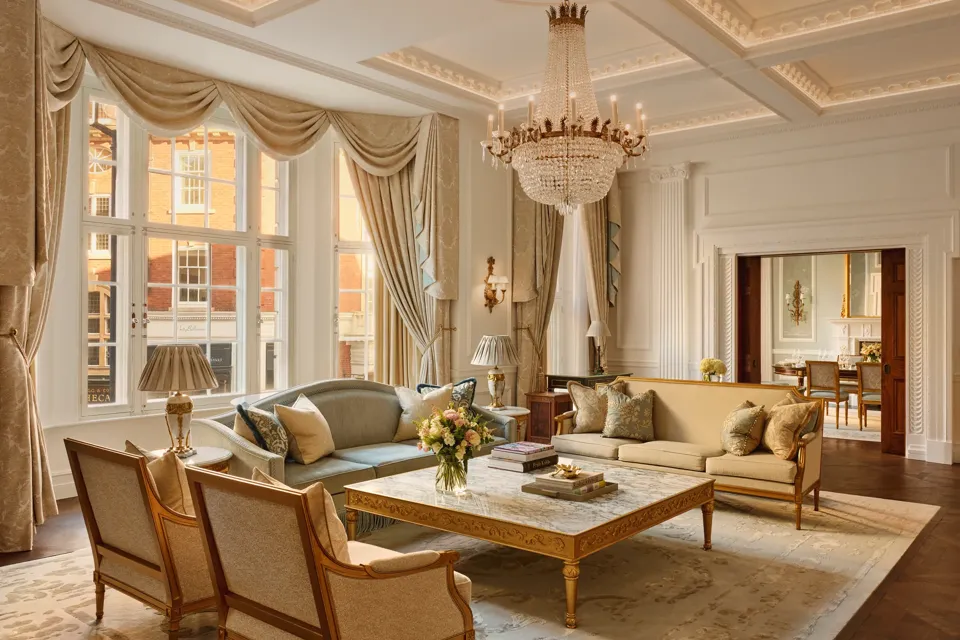 Grand living room with high ceilings, ornate chandelier, and large windows draped in gold curtains, featuring elegant sofas and armchairs around a marble coffee table with carved gold detailing.