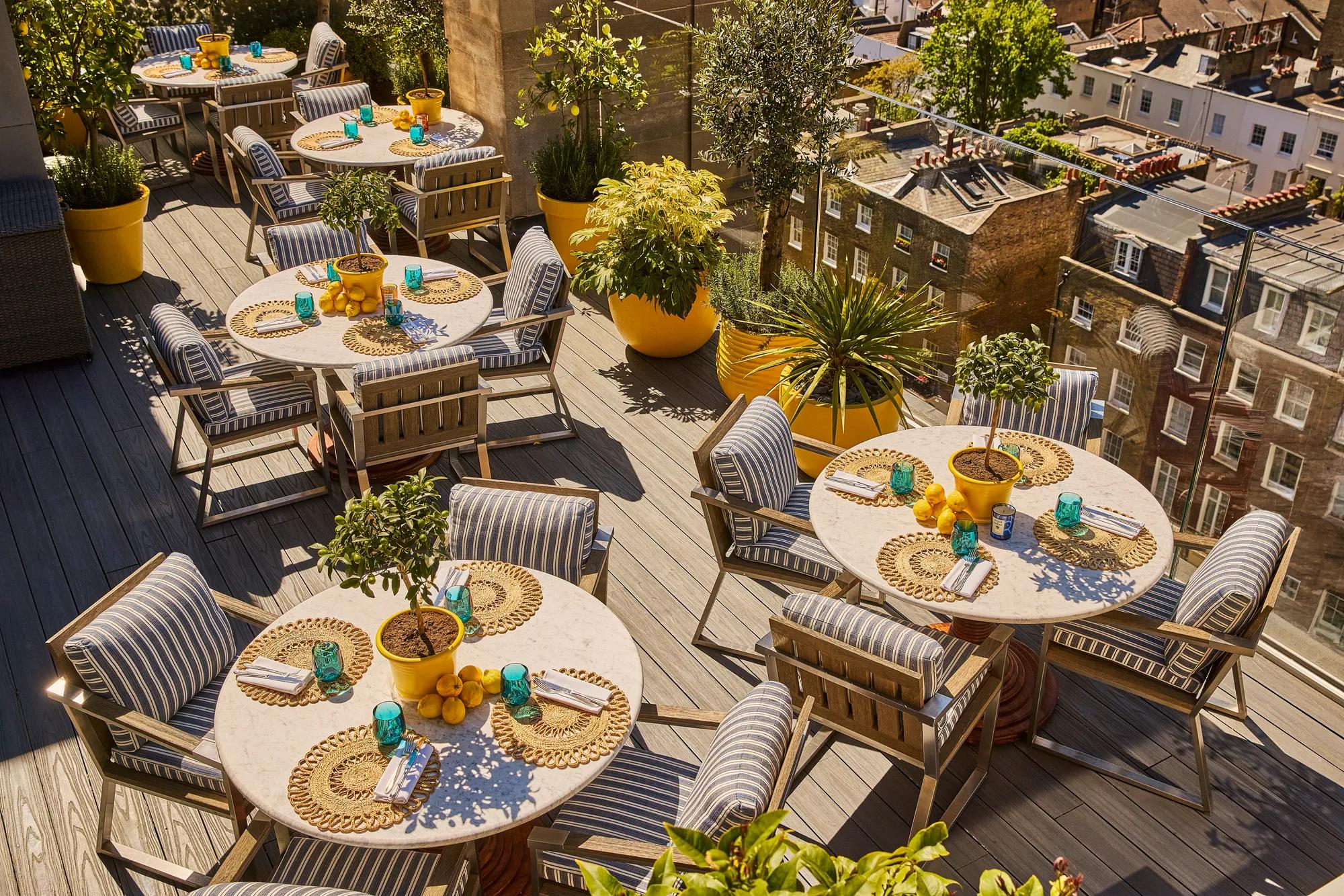 Rooftop terrace dining area with striped chairs, round tables, potted plants and views over surrounding city rooftops.