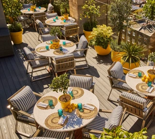 Rooftop terrace dining area with striped chairs, round tables, potted plants and views over surrounding city rooftops.