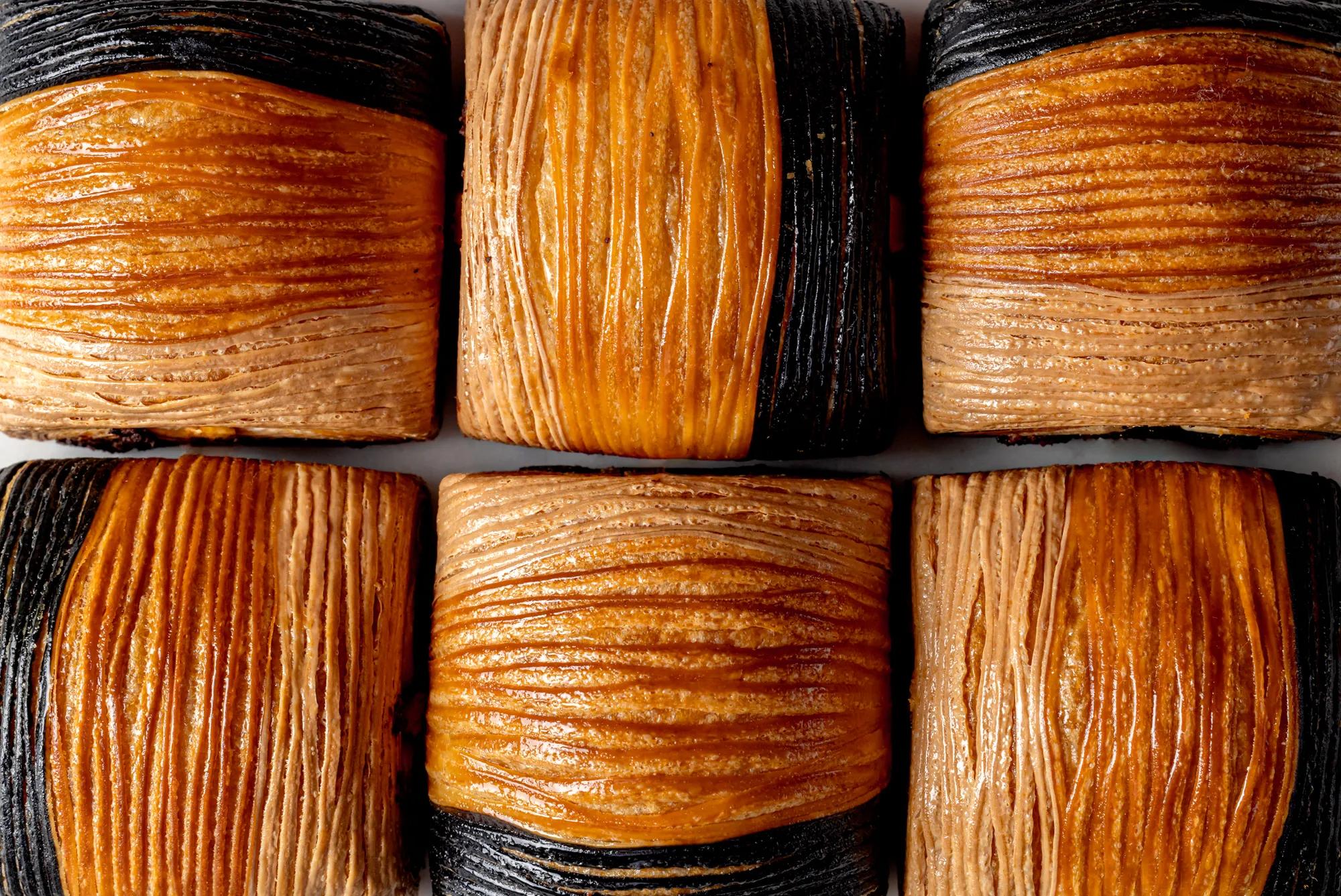 Close-up of flaky croissant pastries with alternating golden brown and dark, charcoal-colored layers in a neat grid.