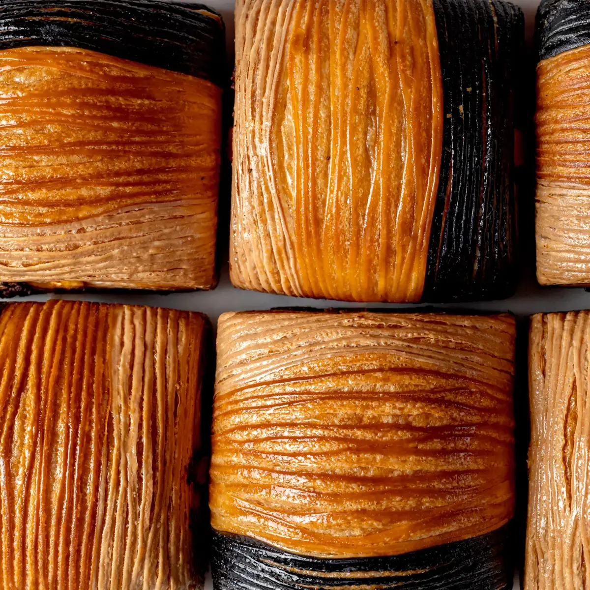 Close-up of flaky croissant pastries with alternating golden brown and dark, charcoal-colored layers in a neat grid.