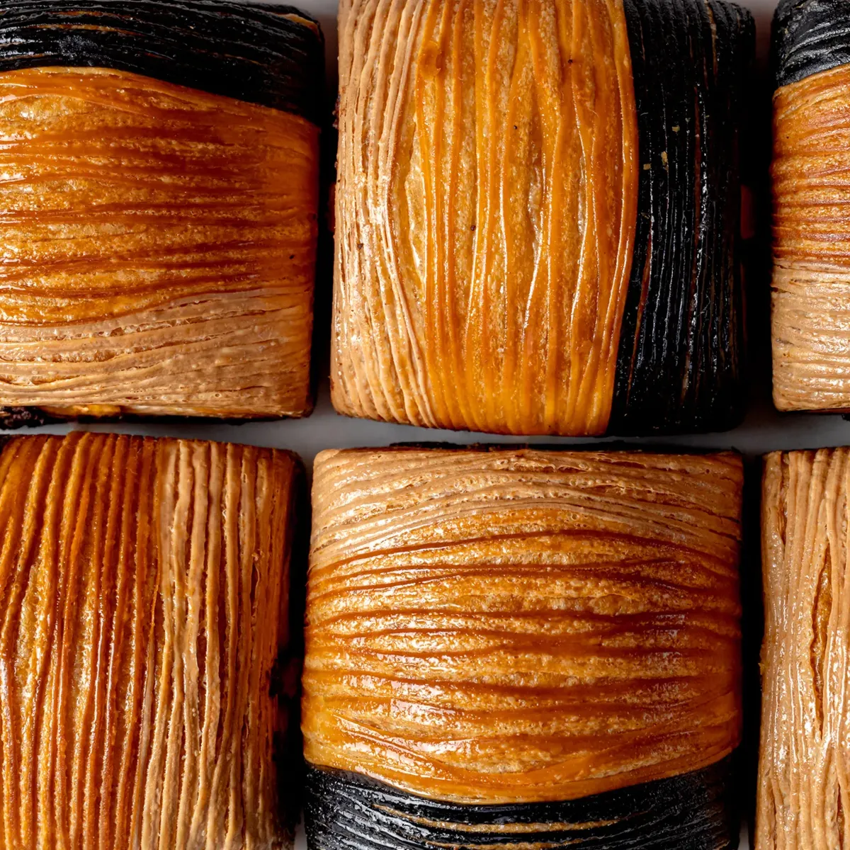 Close-up of flaky croissant pastries with alternating golden brown and dark, charcoal-colored layers in a neat grid.