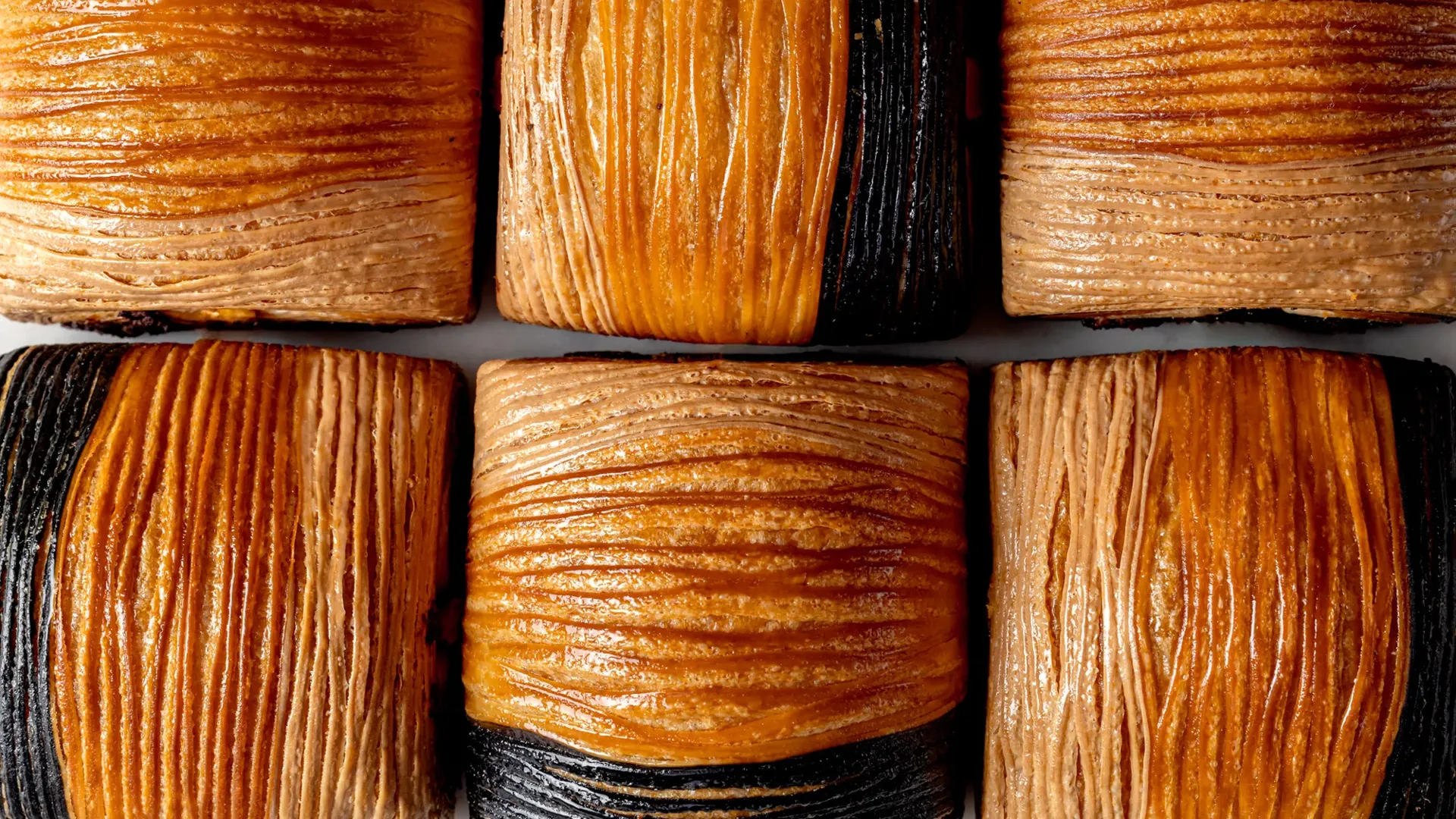 Close-up of flaky croissant pastries with alternating golden brown and dark, charcoal-colored layers in a neat grid.
