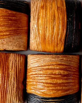 Close-up of flaky croissant pastries with alternating golden brown and dark, charcoal-colored layers in a neat grid.