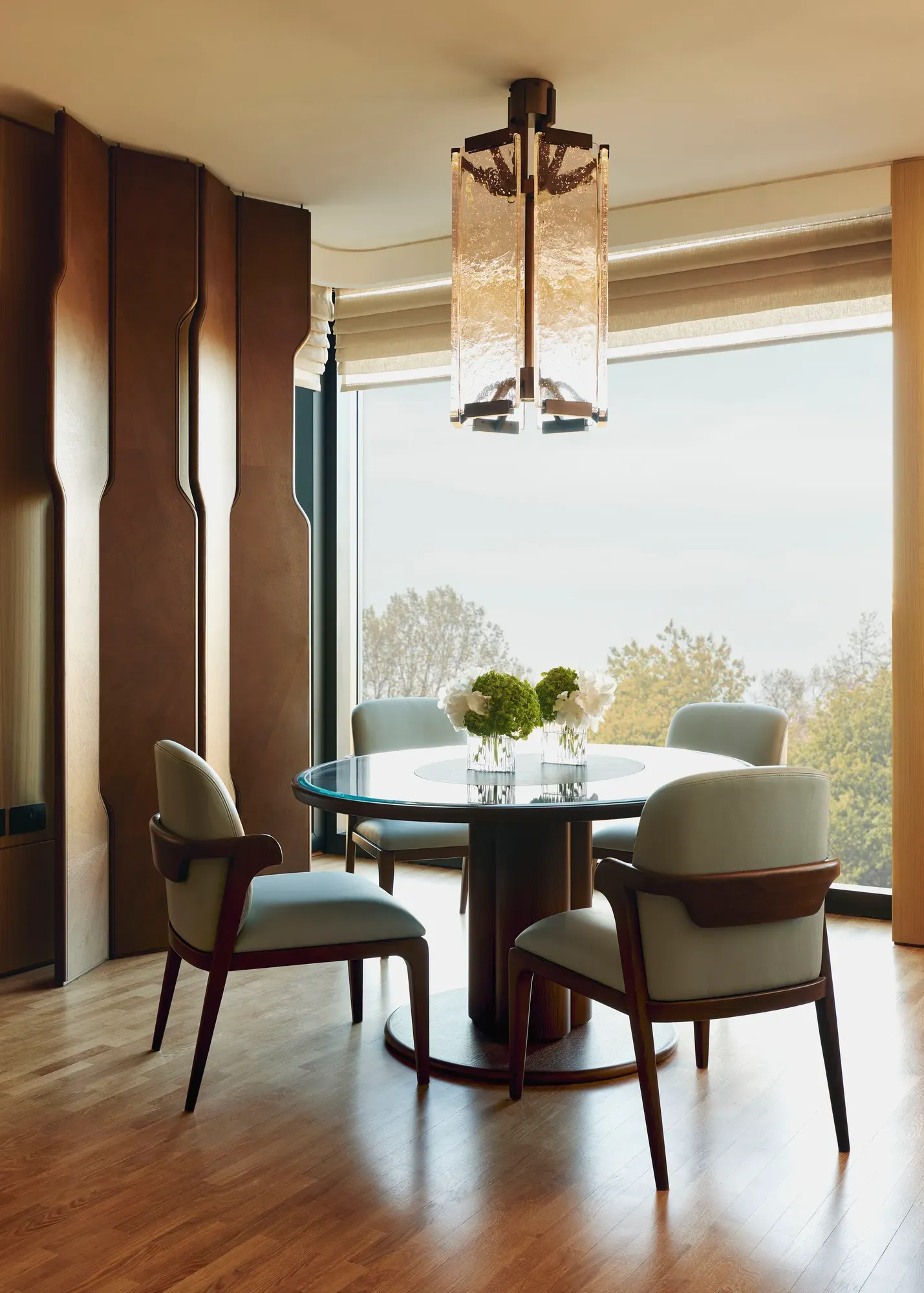 Round dining table with four upholstered chairs, floral centrepiece, large window view of trees, and statement pendant light above.