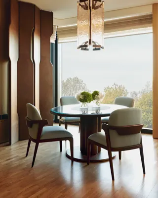 Round dining table with four upholstered chairs, floral centrepiece, large window view of trees, and statement pendant light above.