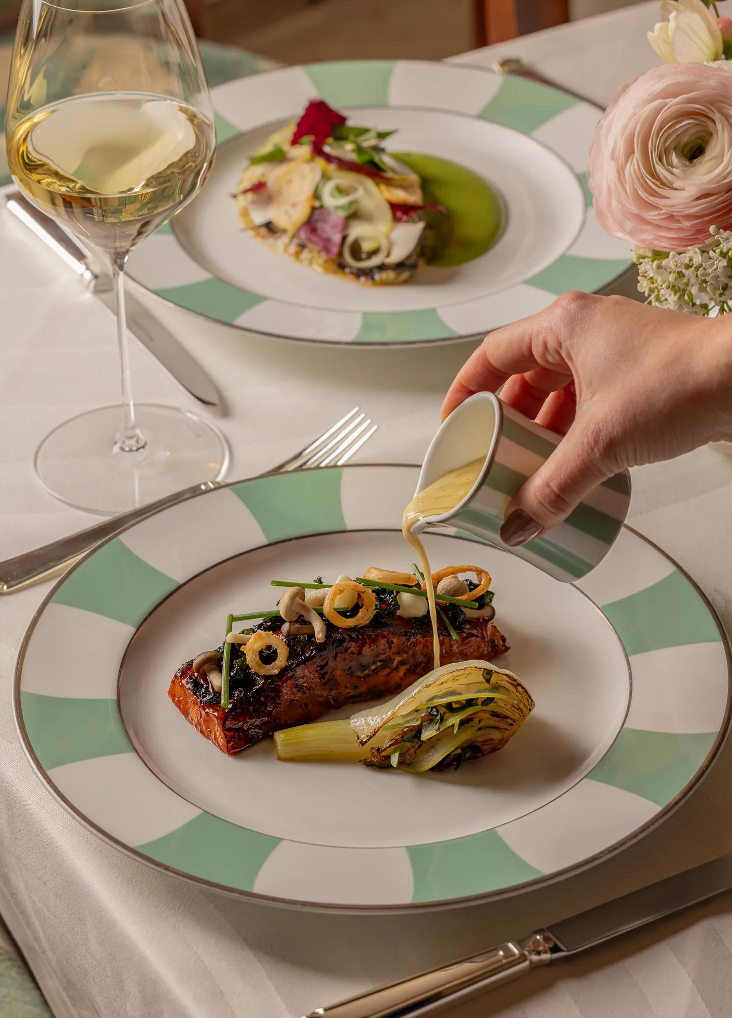 Close-up of a hand pouring sauce over a plated main course of seared fish with grilled vegetables, served on green-and-white patterned china. A second dish, a glass of white wine, and a floral centerpiece complete the elegant table setting.