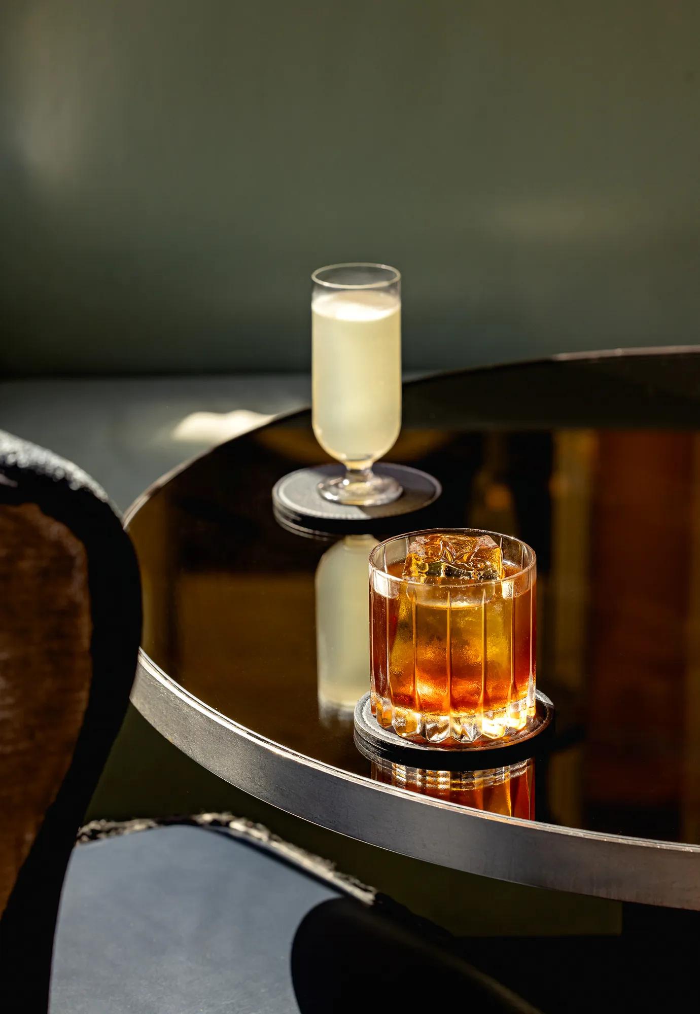 Two cocktails on a glossy round table, one amber in a glass tumbler and one pale drink in a tall stemmed glass.