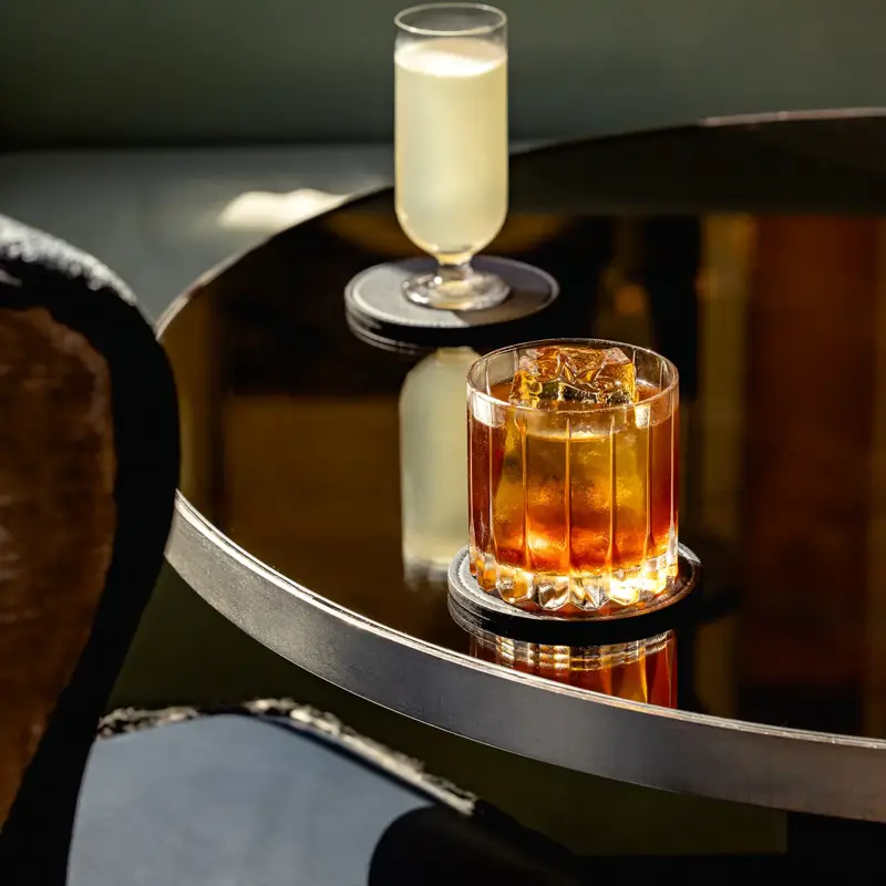 Two cocktails on a glossy round table, one amber in a glass tumbler and one pale drink in a tall stemmed glass.