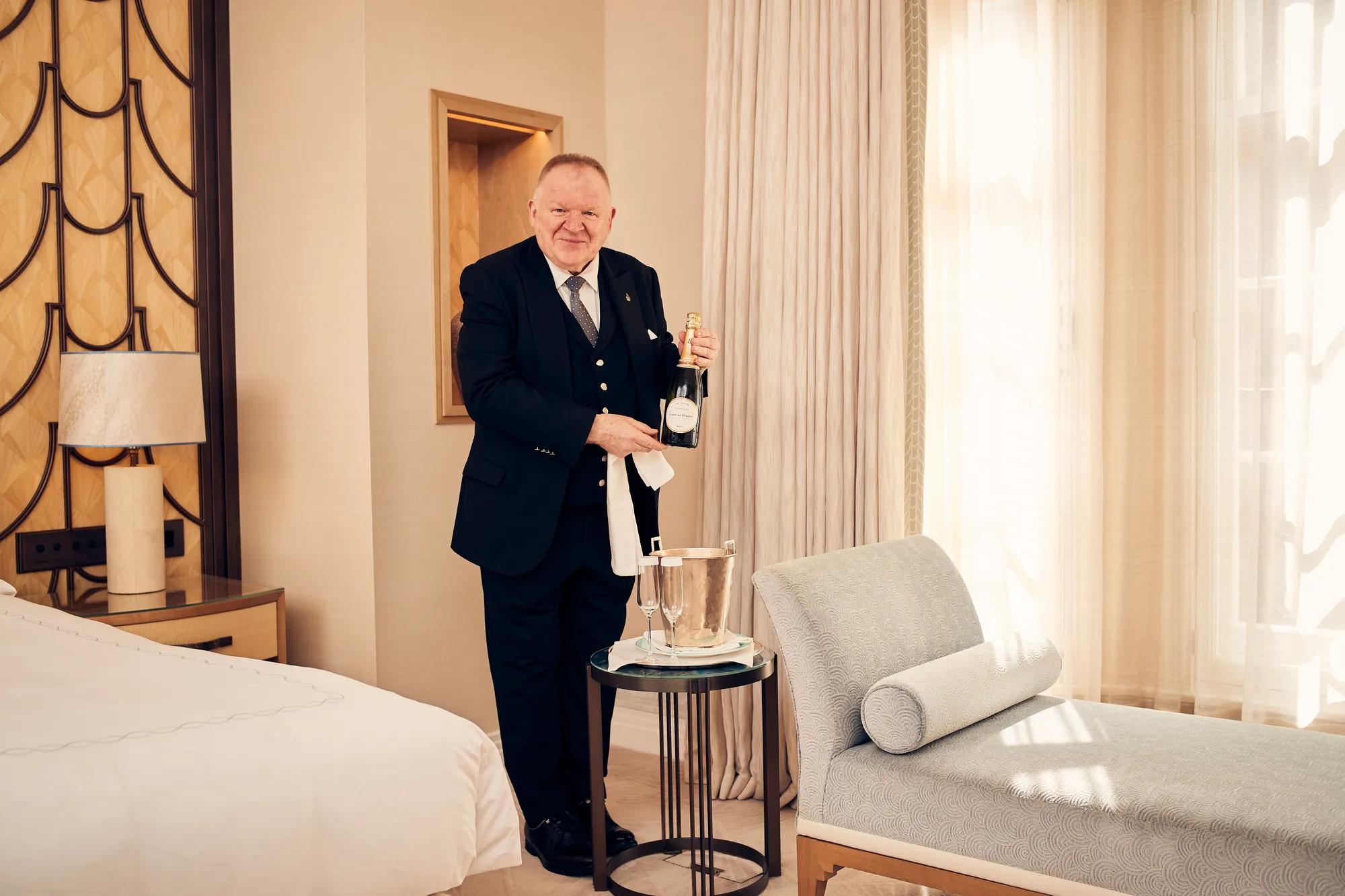 Butler standing in a well-appointed room, holding a bottle of champagne and dressed in formal uniform.